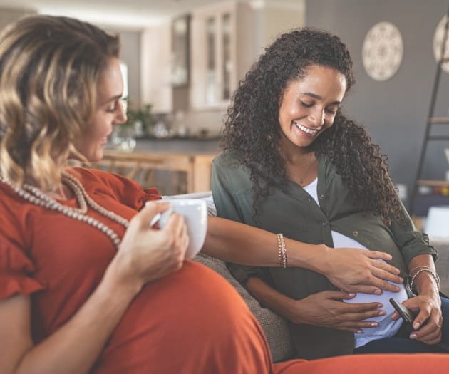 Two pregnant women socialising over a hot beverage. One mother is holding her baby bump and another mother holds her hand out